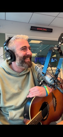 Jon Kamal on air at the radio studio mic wearing headphones, acoustic guitar in the background